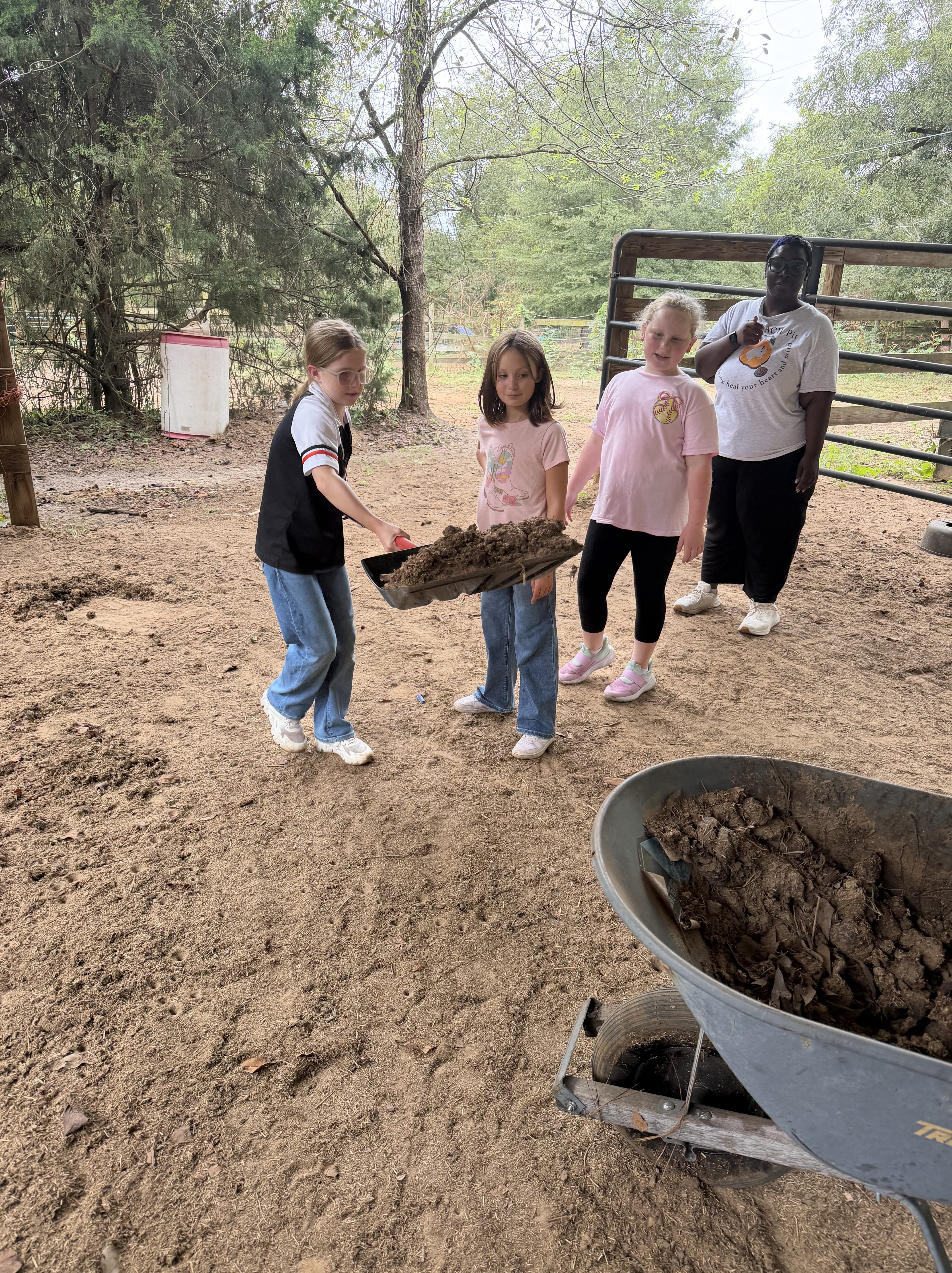 A group of children and an adult outdoors, where one child is holding a shovel with dirt while others watch. A wheelbarrow filled with dirt is in the foreground and trees are visible in the background.