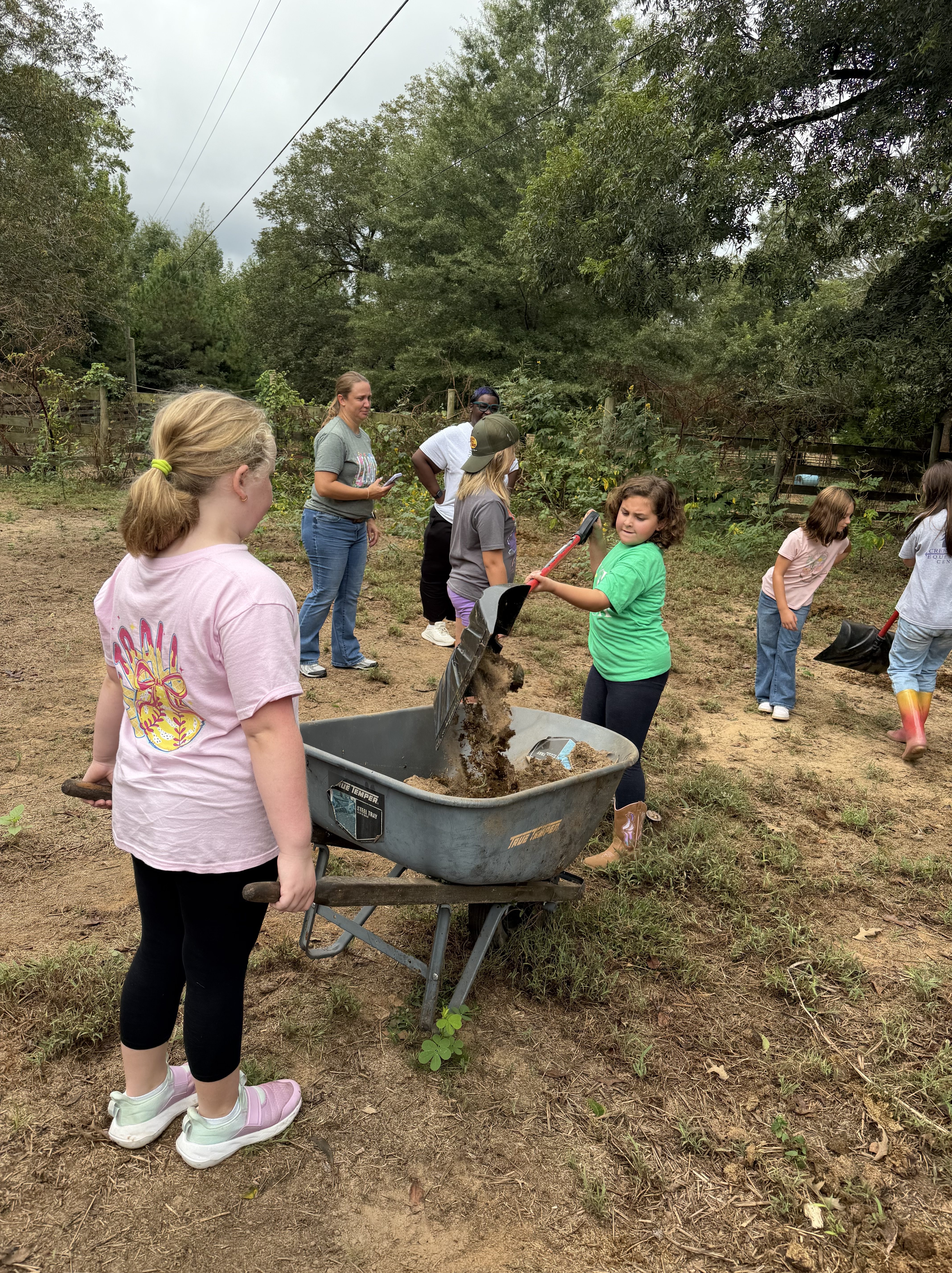 A group of children and adults working together outdoors, with one girl pushing a wheelbarrow filled with soil while others prepare the gardening area in the background.