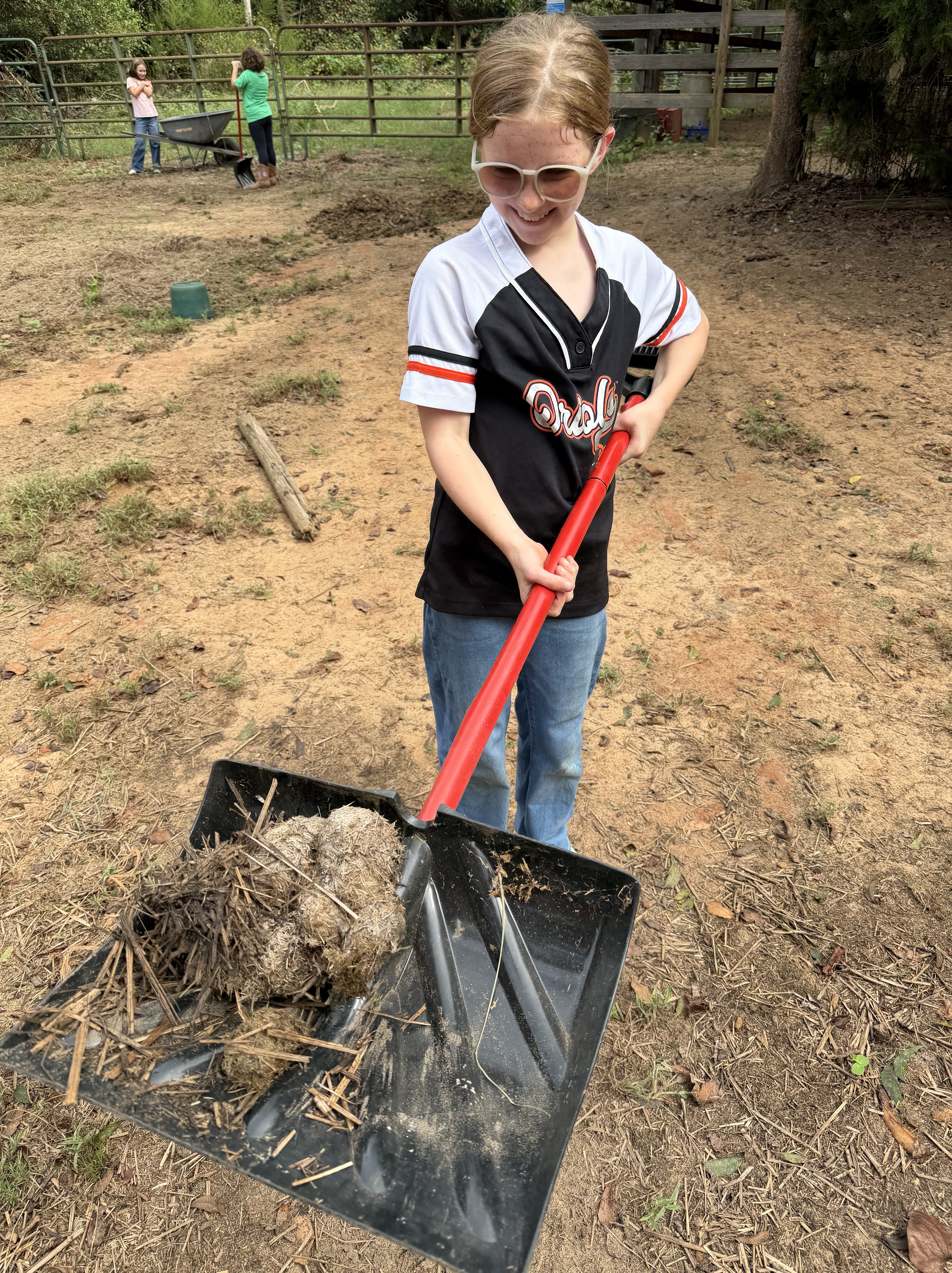 A child with glasses holds a shovel filled with dirt and debris on a farm, while other children are seen in the background.