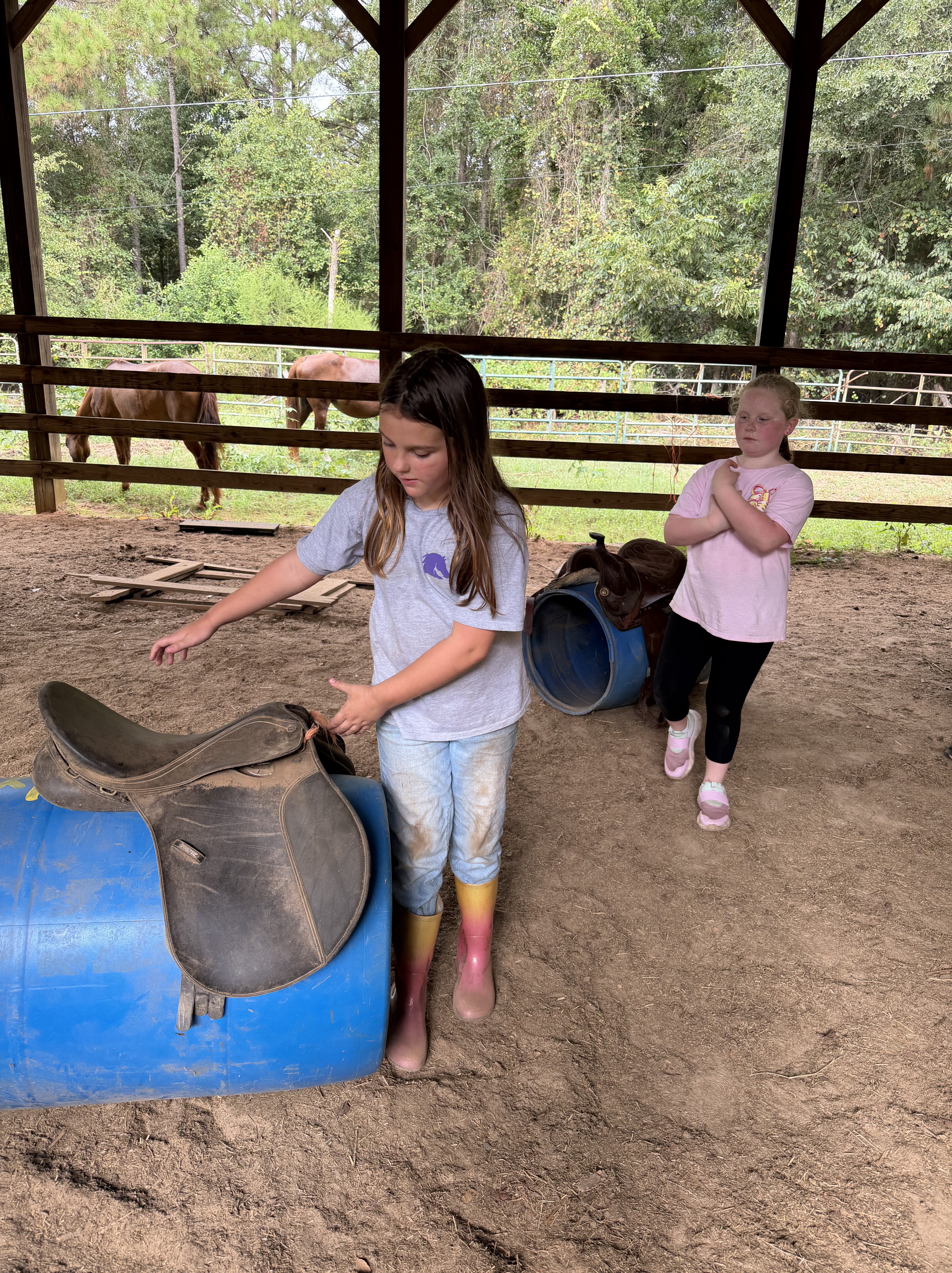 Two children in a barn setting, one is arranging a saddle on a blue barrel, while the other stands nearby with arms crossed. In the background, horses can be seen grazing.